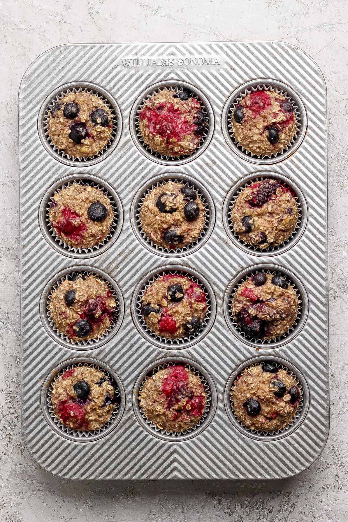 A muffin tin filled with twelve unbaked oat muffins, topped with blueberries and raspberries, on a light-colored surface.