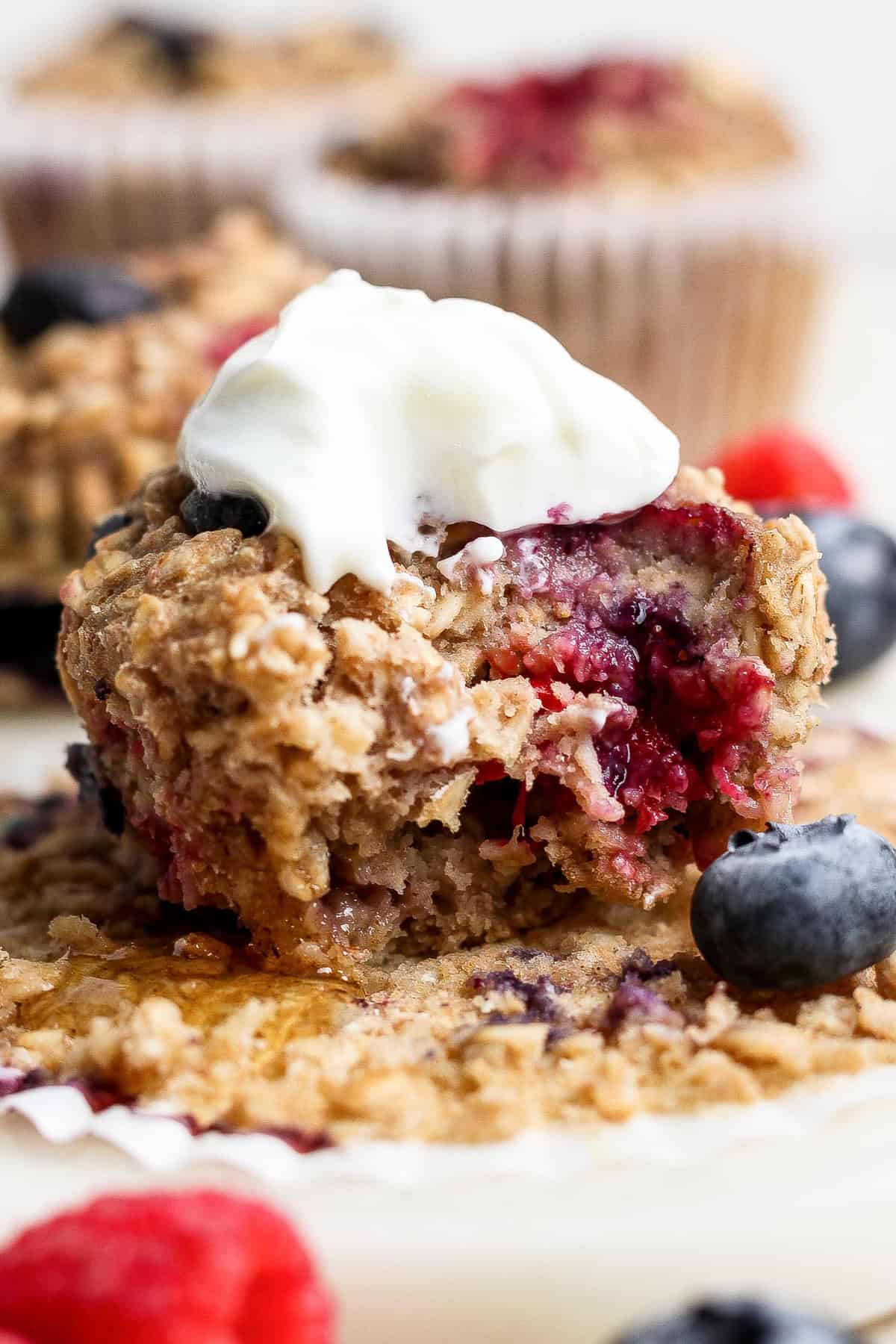 Get your meal prep on and make these easy Triple Berry Baked Oatmeal Cups for the week's breakfast. This baked oatmeal recipe is made with 3 kinds of berries, rolled oats, and ground oat flour making this a better-for-you breakfast or snack! A close-up of a berry muffin with a bite taken out, topped with a dollop of cream, surrounded by fresh blueberries and raspberries.