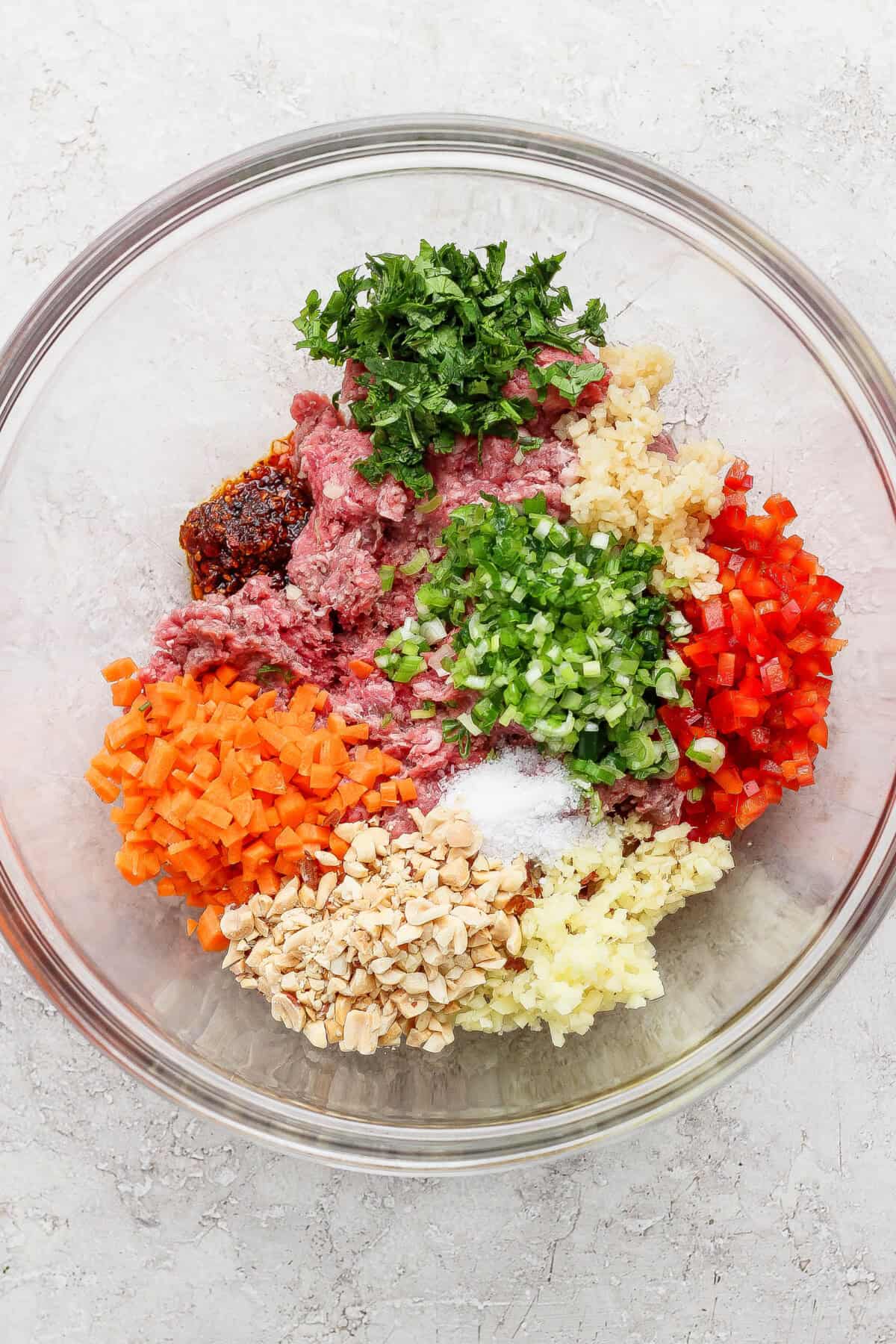 A glass bowl containing ground meat, chopped carrots, bell peppers, mushrooms, parsley, ginger, garlic, green onions, and seasonings arranged in separate piles.