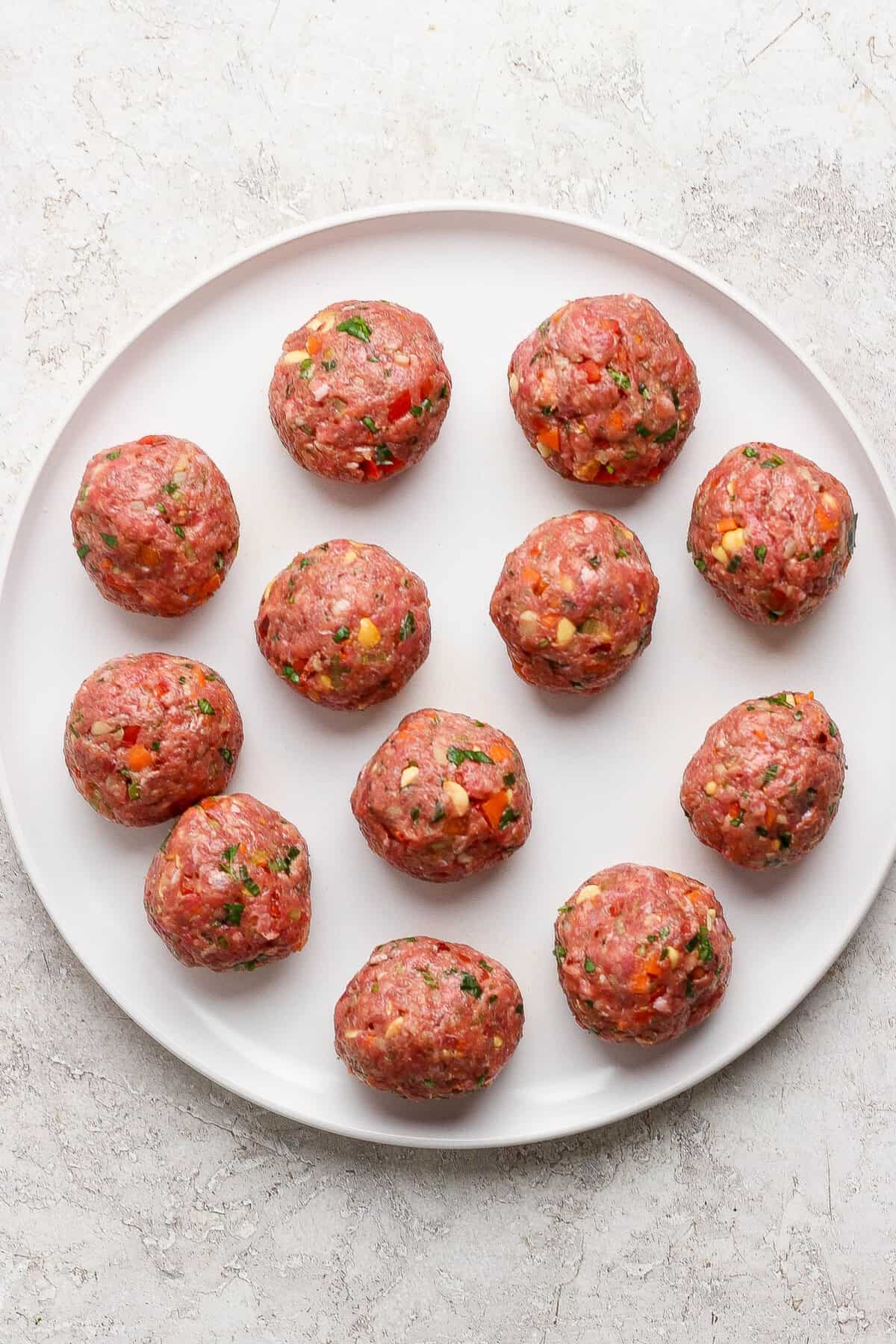 A white plate with twelve uncooked meatballs, mixed with herbs and small vegetable pieces, on a light textured surface.