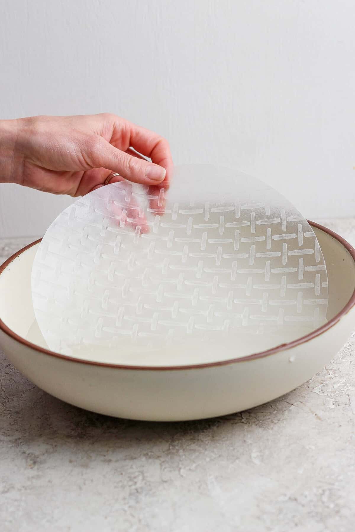 A hand dips a round sheet of rice paper into a shallow bowl of water on a light-colored surface.