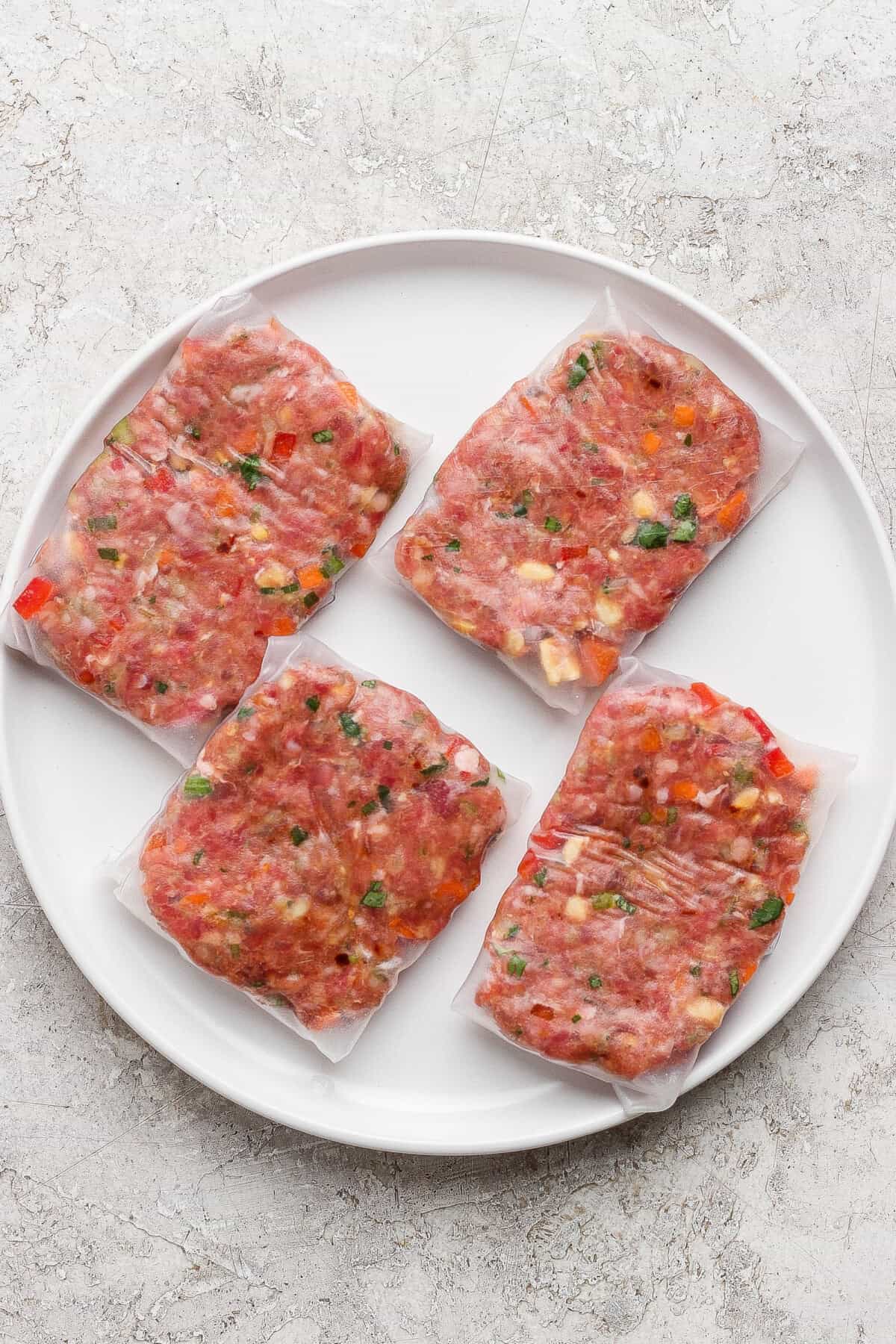 Four vacuum-sealed portions of seasoned raw ground meat with visible herbs and vegetables, arranged on a round white plate.