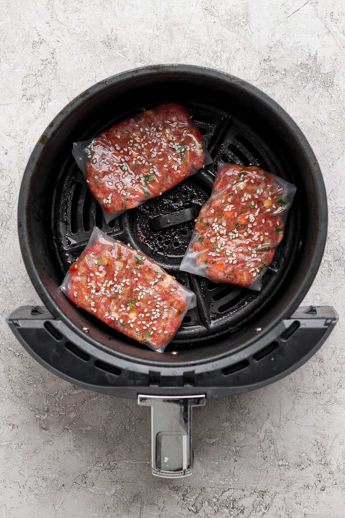 Three uncooked frozen salmon filets coated with marinade and sesame seeds arranged in the basket of an air fryer.