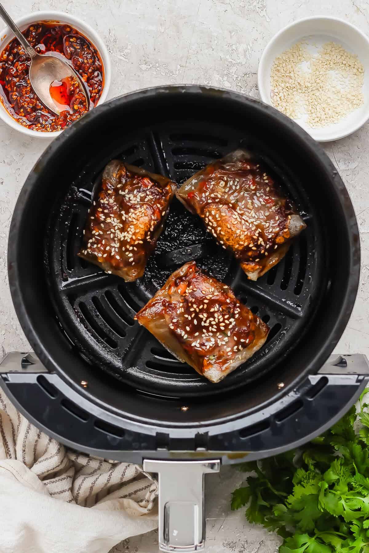 Three pieces of marinated fish with sesame seeds in an air fryer basket, with a bowl of chili sauce and a bowl of sesame seeds nearby.