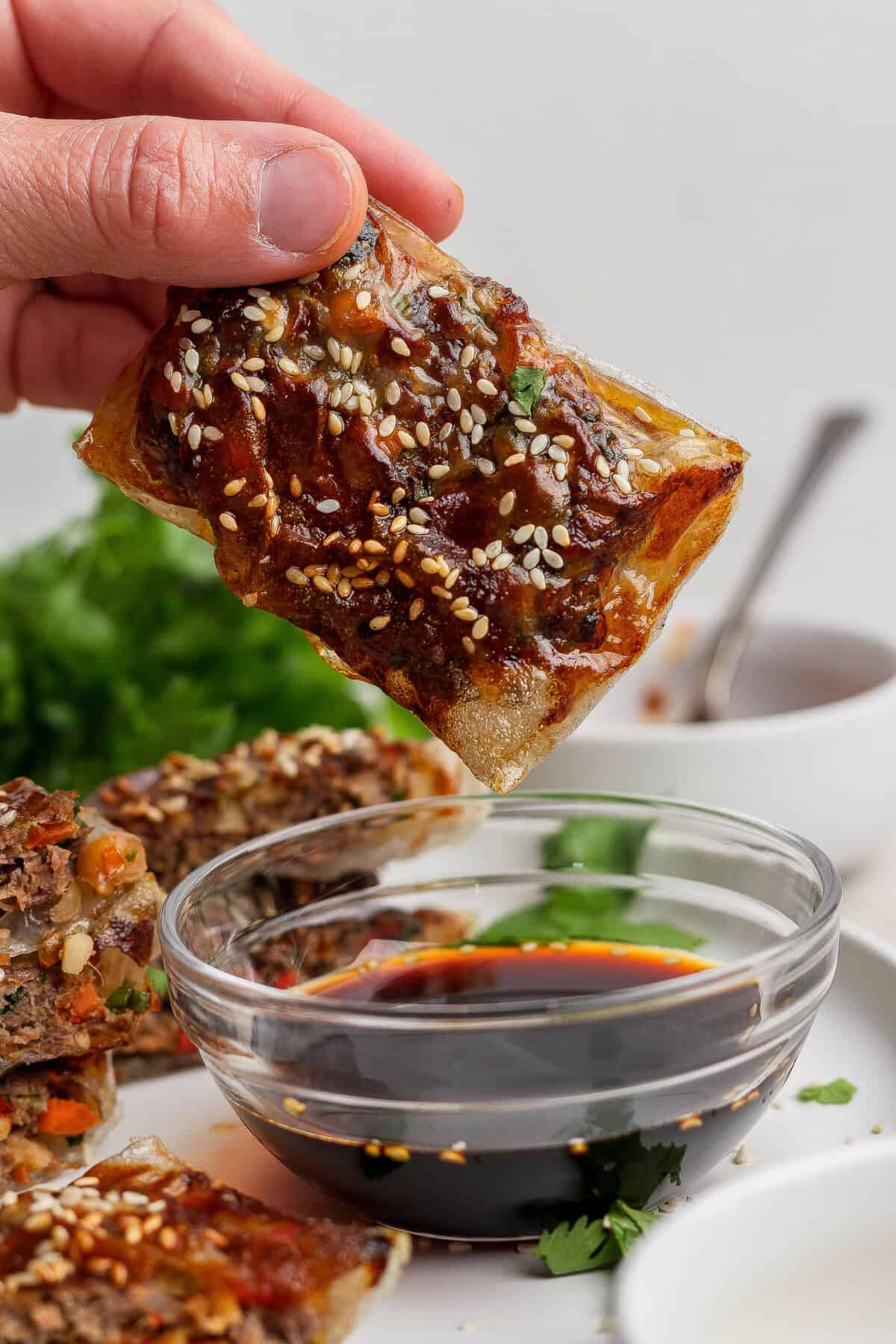 A hand holds a crispy, rectangular pastry topped with sesame seeds, about to dip it into a bowl of soy sauce; greens and more pastries are in the background.