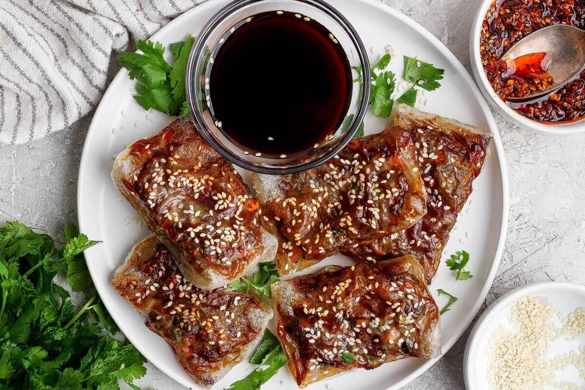 A white plate with five pieces of crispy, glazed tofu topped with sesame seeds, garnished with cilantro, and served with a small bowl of dark dipping sauce.