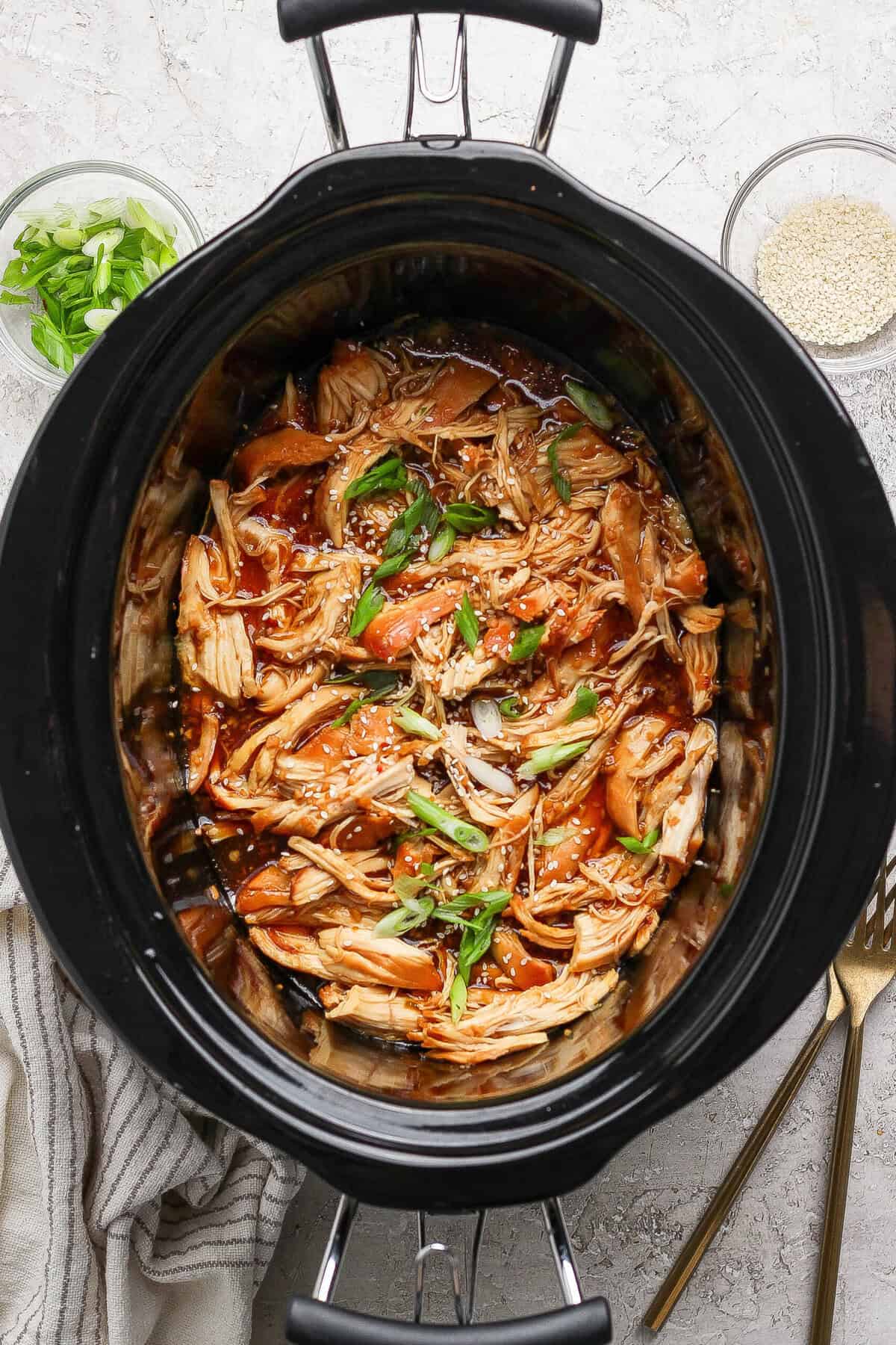 Shredded crockpot honey garlic chicken topped with green onions and sesame seeds in a slow cooker