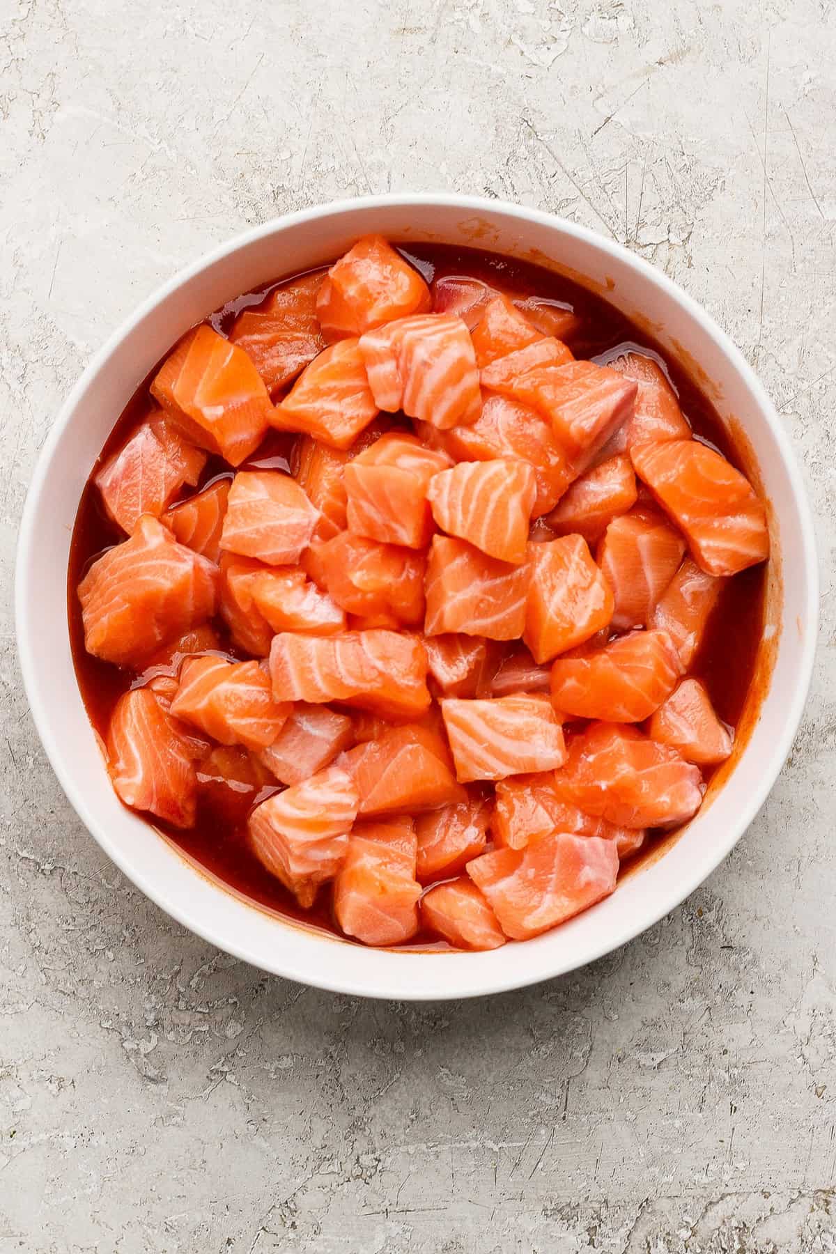 A white bowl filled with cubed raw salmon mixed with a red marinade, placed on a light textured surface.