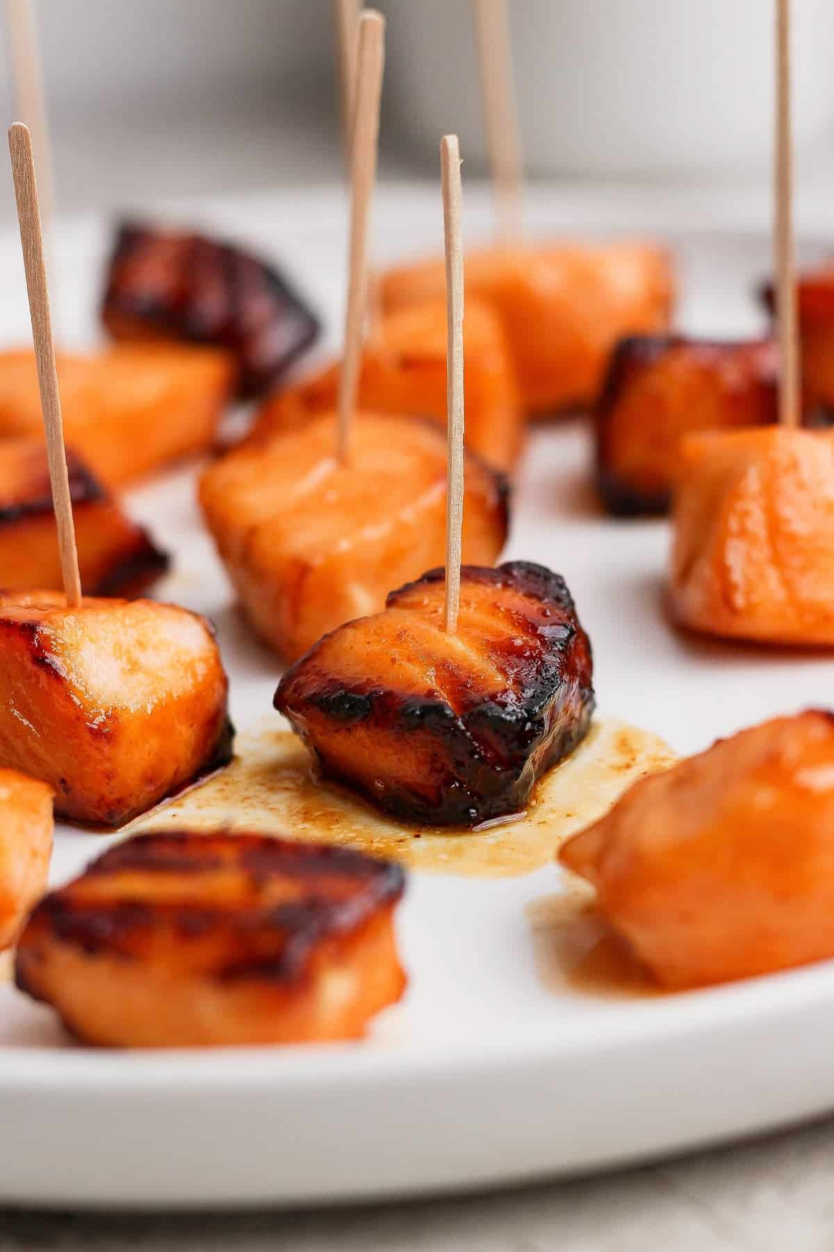 Cubed pieces of glazed salmon, some with char marks, arranged on a white plate and skewered with wooden toothpicks.