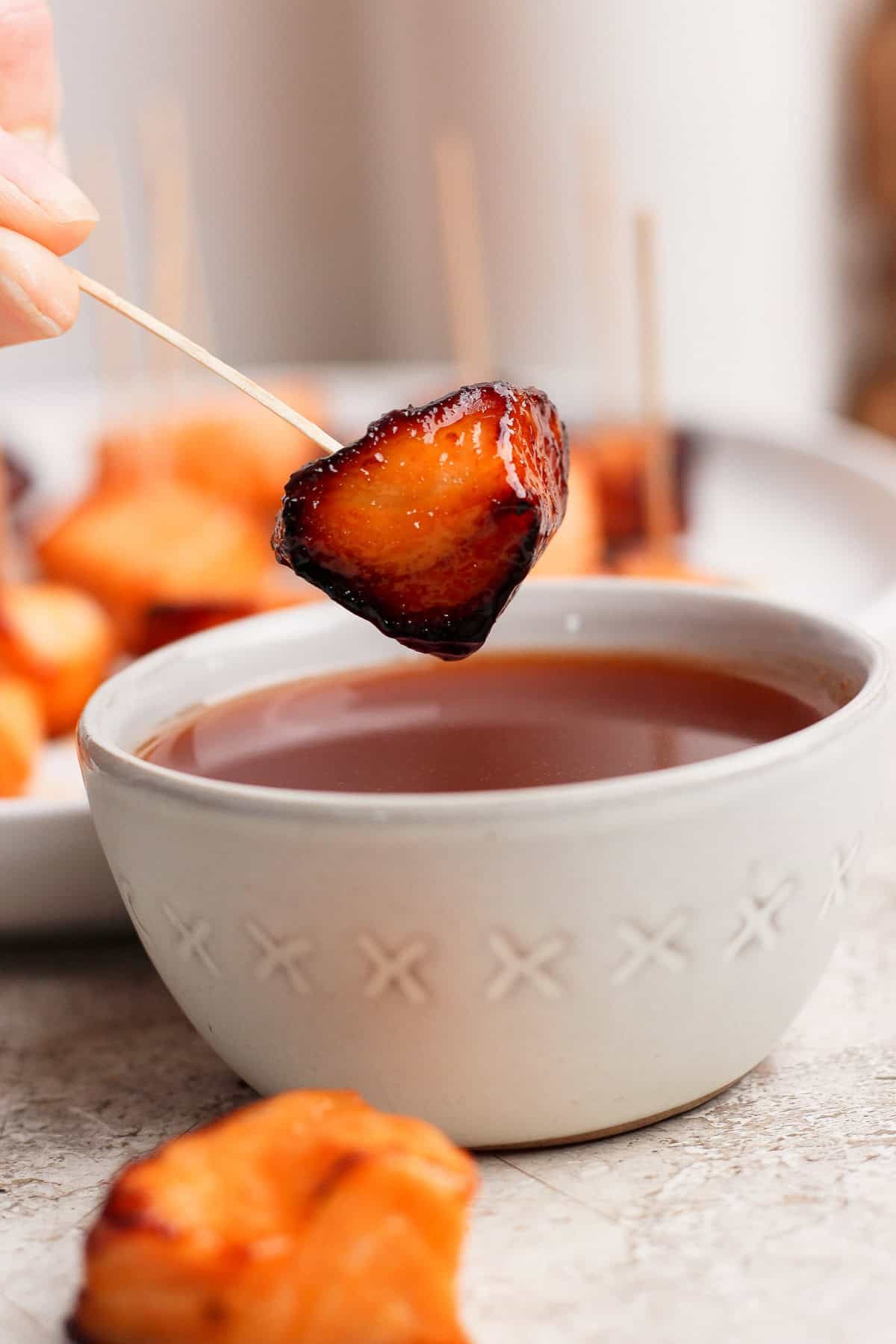 A hand holds a skewer with a glazed, roasted bite-sized food piece above a bowl of dipping sauce; more skewered pieces are seen in the background.