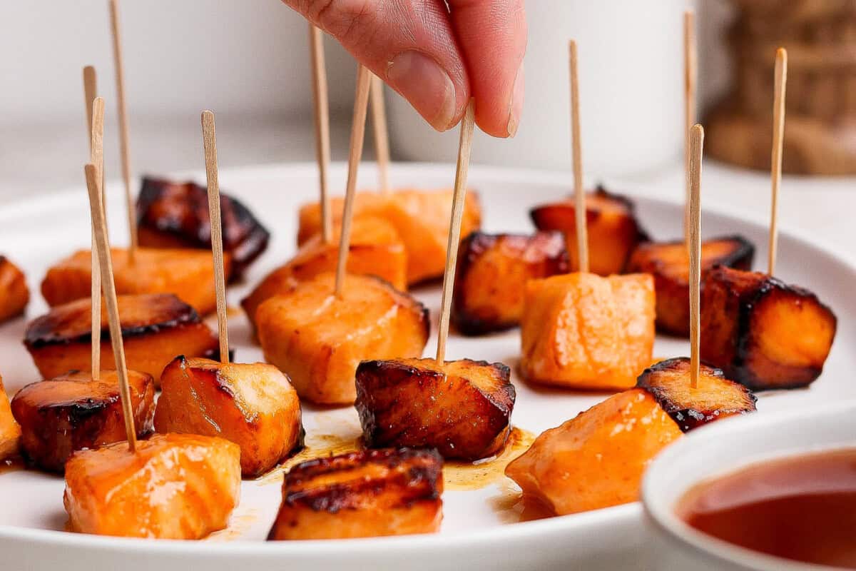 A hand picks up a toothpick with a piece of roasted sweet potato from a plate filled with several other sweet potato bites on toothpicks.