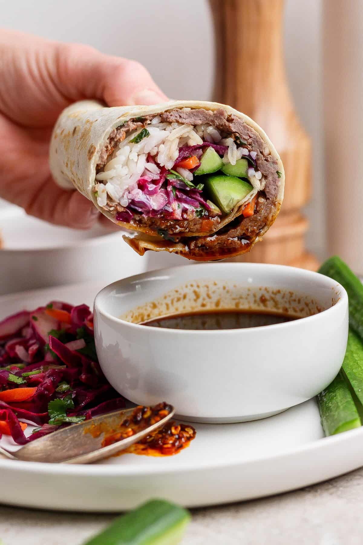 A hand holds a partially eaten wrap filled with rice, vegetables, and sauce above a white bowl of dipping sauce on a plate with salad and cucumber slices.