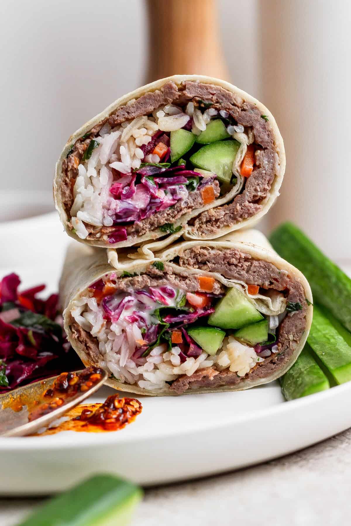 A close-up of a halved wrap filled with rice, vegetables, and a ground meat mixture, served on a white plate with cucumber slices and a side of spicy sauce.