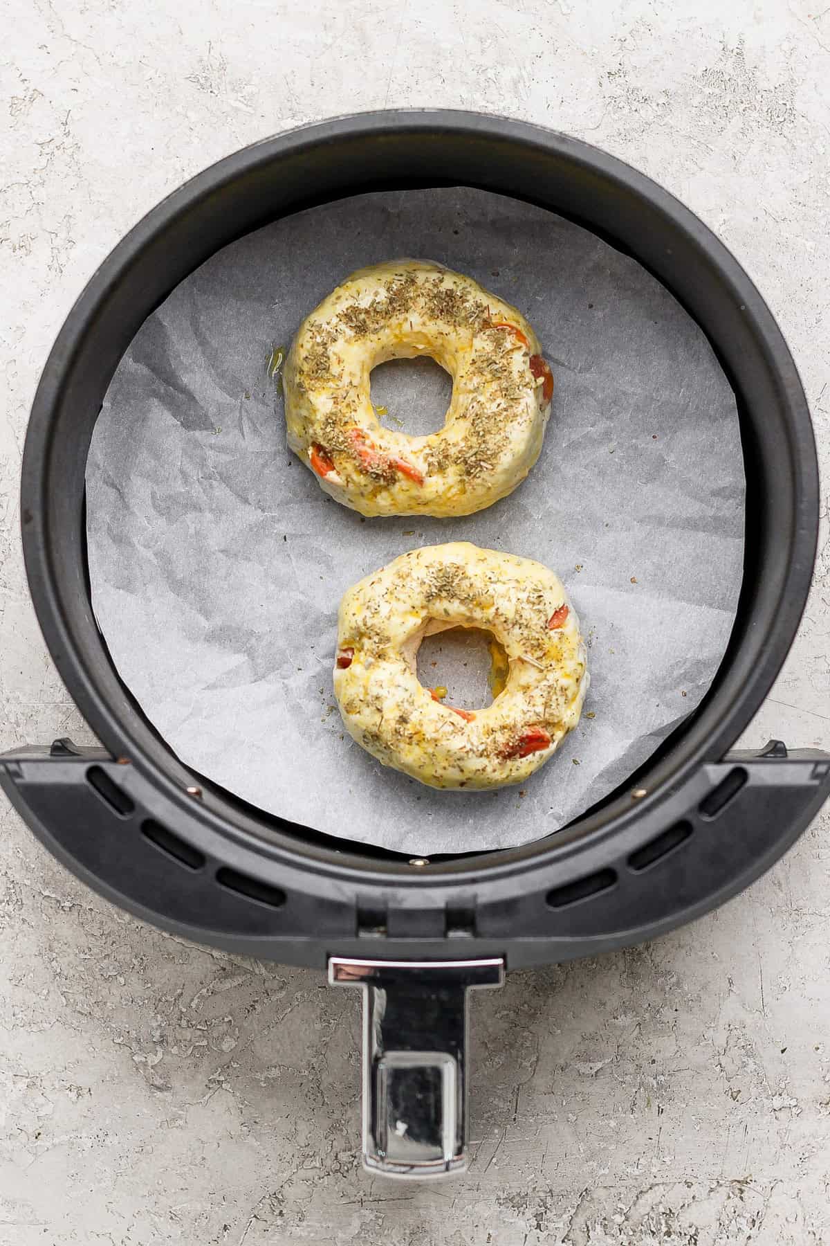 Two uncooked pizza protein bagels with seasoning in an air fryer basket