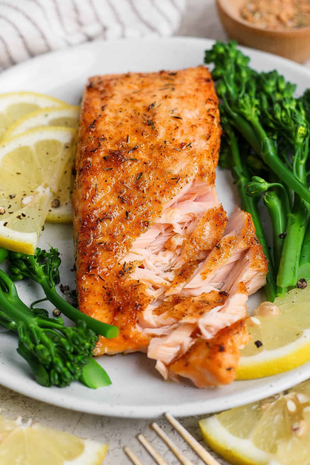Flaky broiled salmon fillet on a white plate with broccolini and lemon, showing the tender interior.