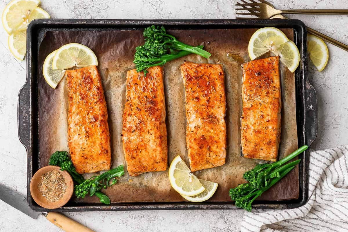 Broiled salmon fillets on a baking sheet garnished with lemon slices, broccolini, and a bowl of seasoning.