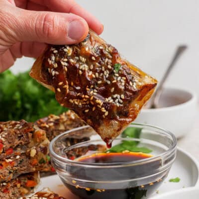 A hand dips a rectangular, sesame seed-topped pastry into a bowl of dark soy sauce. Other similar pastries and green herbs are visible in the background.