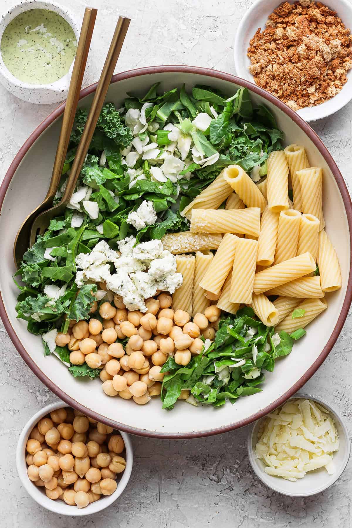 A large bowl with rigatoni pasta, chickpeas, leafy greens, feta cheese, and shaved parmesan, surrounded by bowls of dressing, crumbled topping, extra chickpeas, and parmesan.