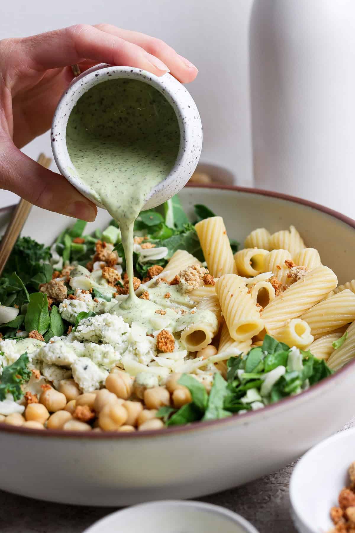 A hand pours creamy green dressing over a bowl of pasta salad with rigatoni, chickpeas, leafy greens, and crumbled toppings.