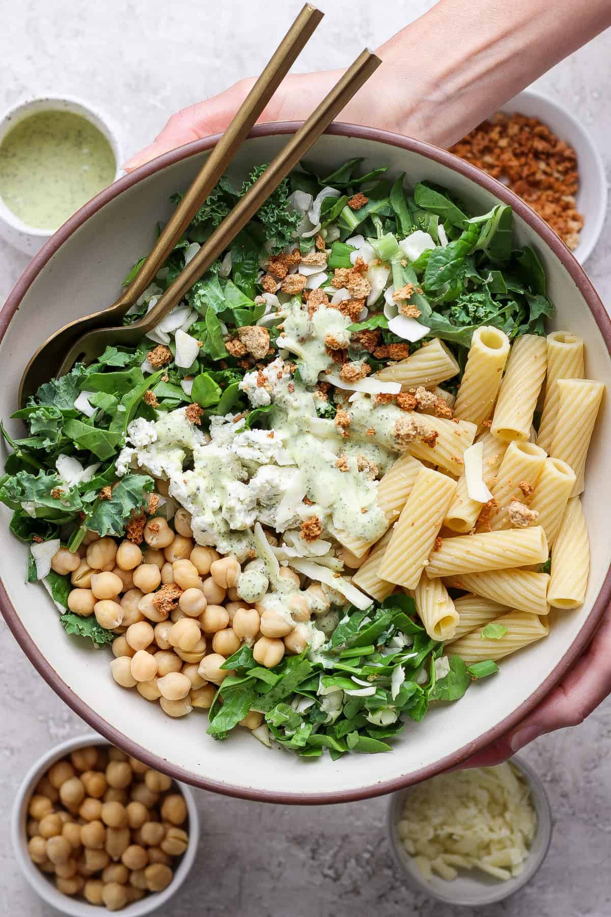 A bowl containing mixed greens, rigatoni pasta, chickpeas, cheese, and a creamy dressing, with two forks. Surrounding bowls hold extra chickpeas, shredded cheese, dressing, and crunchy toppings.