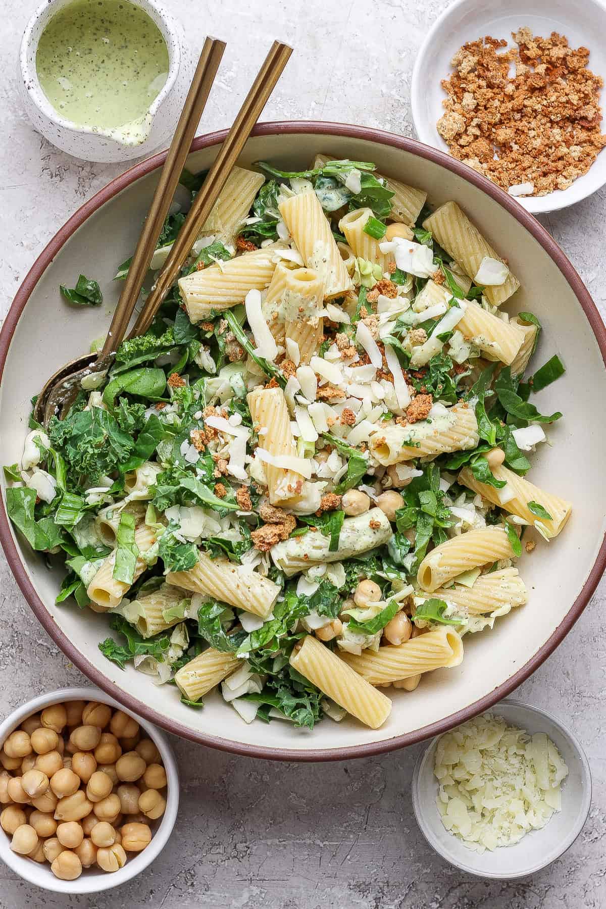 A bowl of pasta salad with greens, chickpeas, cheese, and crumb topping, surrounded by small bowls of chickpeas, cheese, crumbs, and dressing.