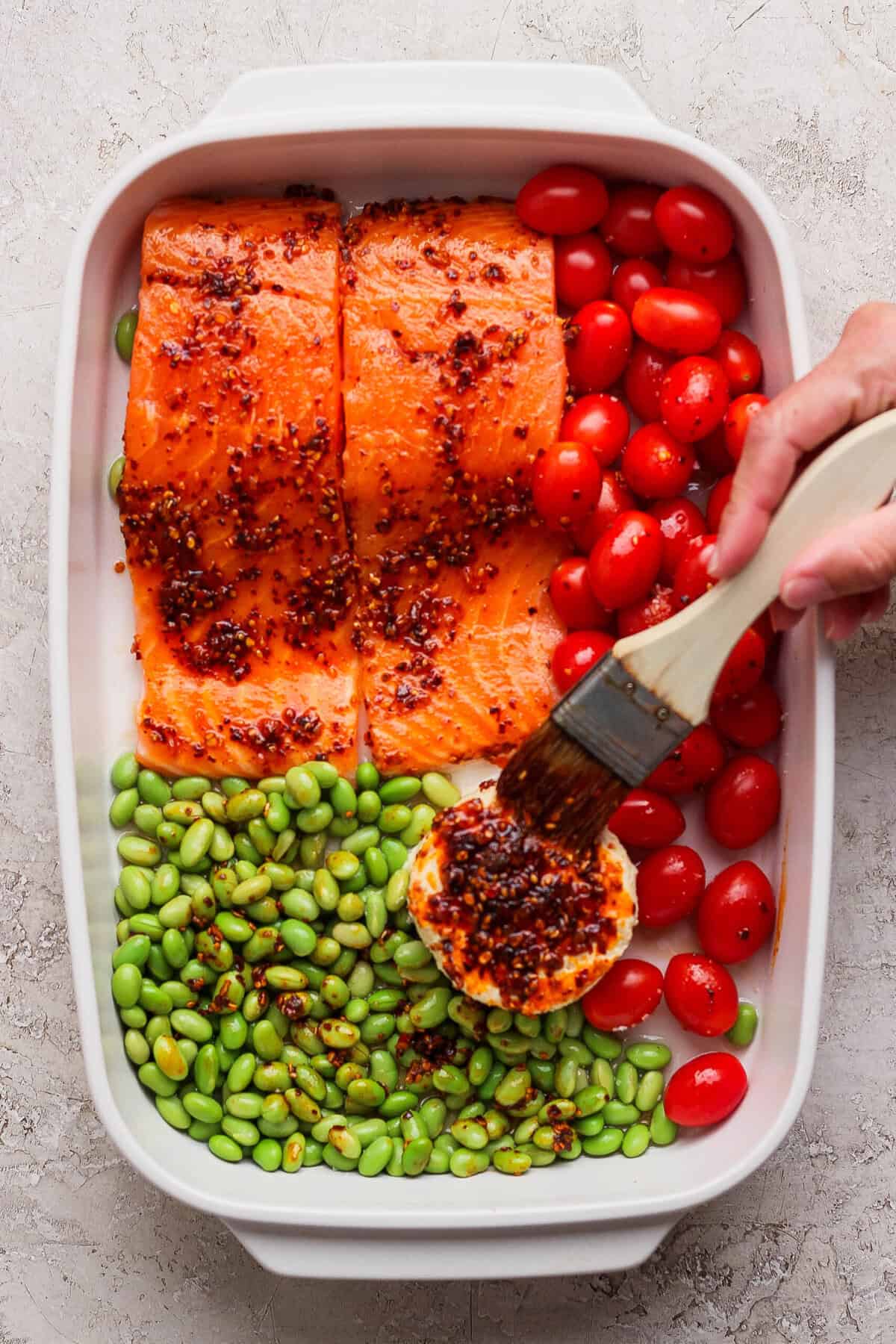 Brushing chili crunch onto Boursin cheese in a casserole dish with raw salmon and vegetables