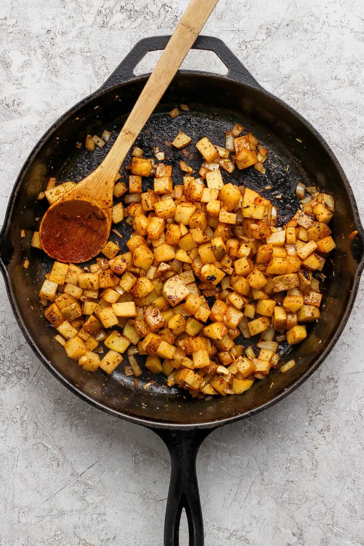 Diced potatoes and onions seasoned and cooked in a cast iron skillet with a wooden spoon on top.