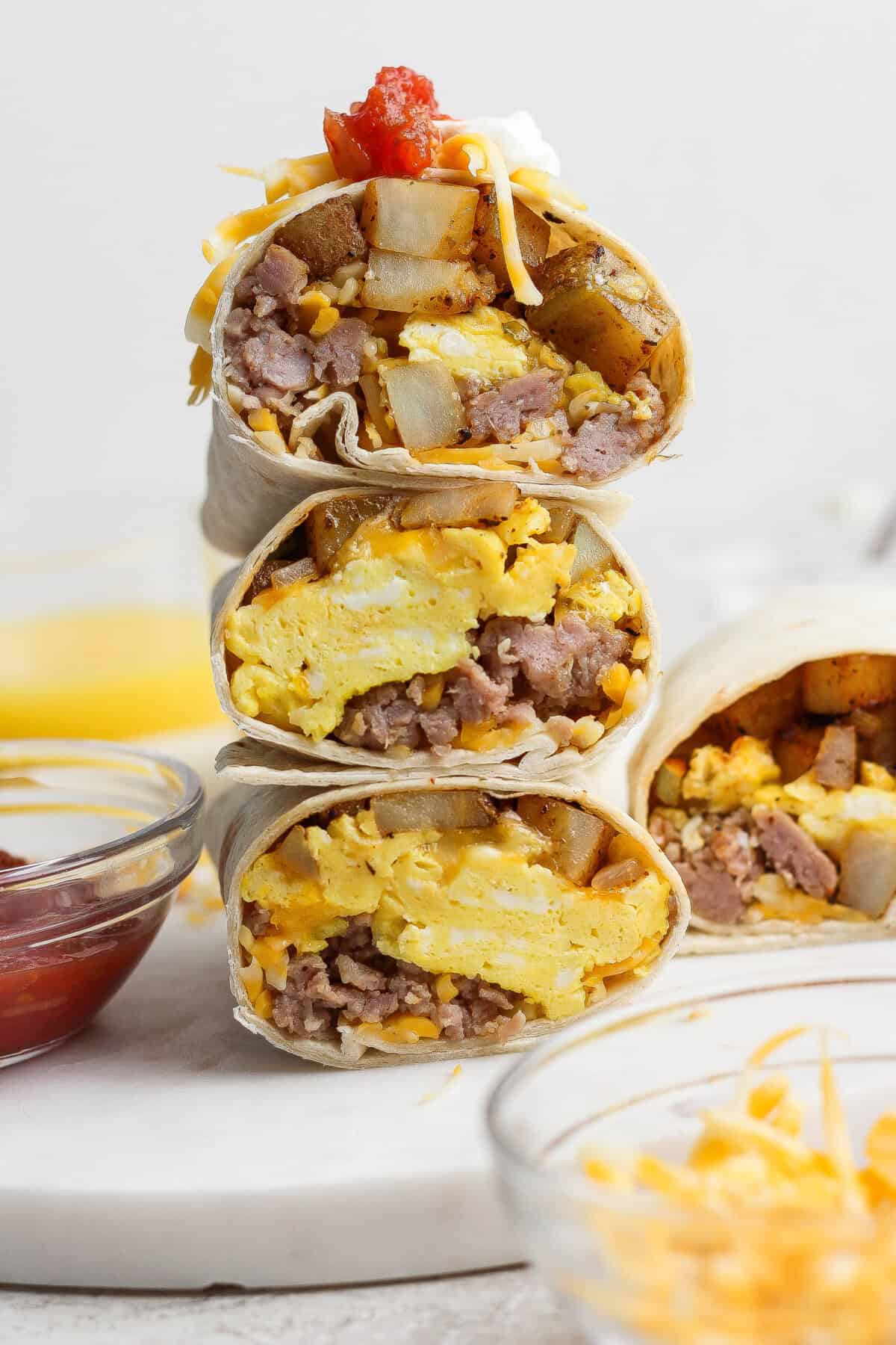 These make ahead breakfast burritos are the perfect grab-n-go breakfast choice for your busy mornings. They are packed with veggies and protein to keep your energy up all day. Make a double batch of these freezer breakfast burritos for an easy, healthy meal-prep breakfast you can enjoy all week long. Three breakfast burritos cut in half and stacked, showing scrambled eggs, sausage, potatoes, and cheese inside; surrounded by bowls of ketchup, shredded cheese, and orange juice.