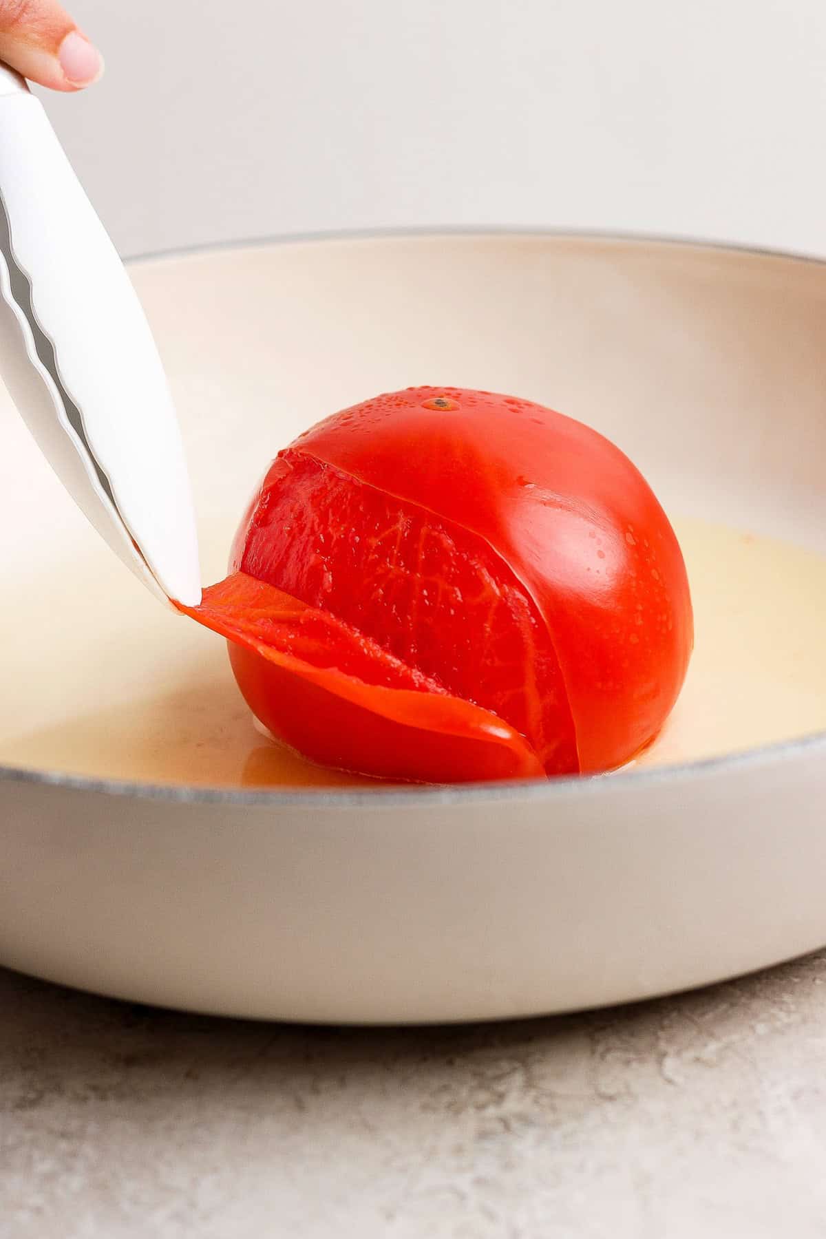 Tongs removing the loosened peel from a steamed whole tomato in a skillet.