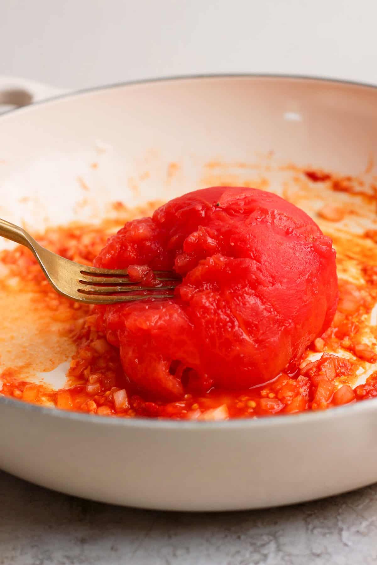 A peeled steamed tomato being smashed into onion, garlic, and tomato paste with a fork.