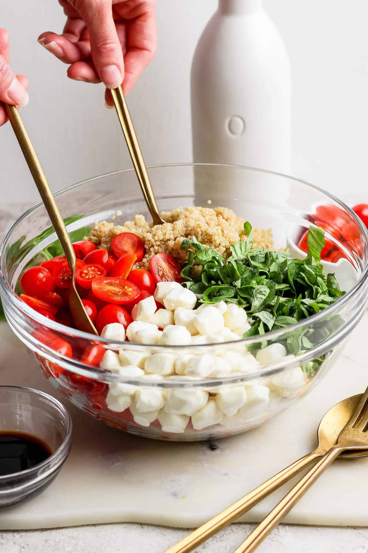 A person tosses a salad in a glass bowl with mozzarella balls, cherry tomatoes, quinoa, spinach, and basil. A bottle and small bowl of balsamic vinegar are nearby.