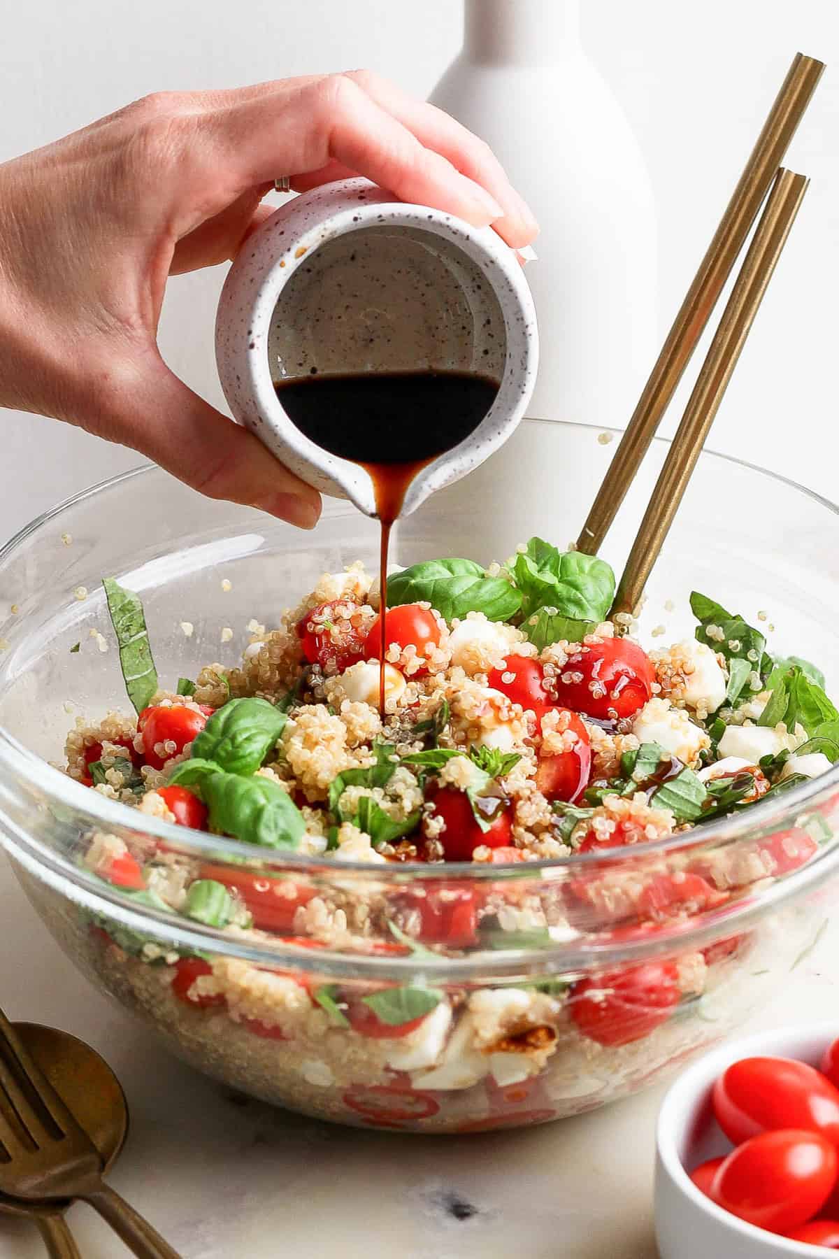 Hand pouring dark dressing onto a quinoa salad with cherry tomatoes, basil, and mozzarella in a glass bowl, with chopsticks and a white vase in the background.