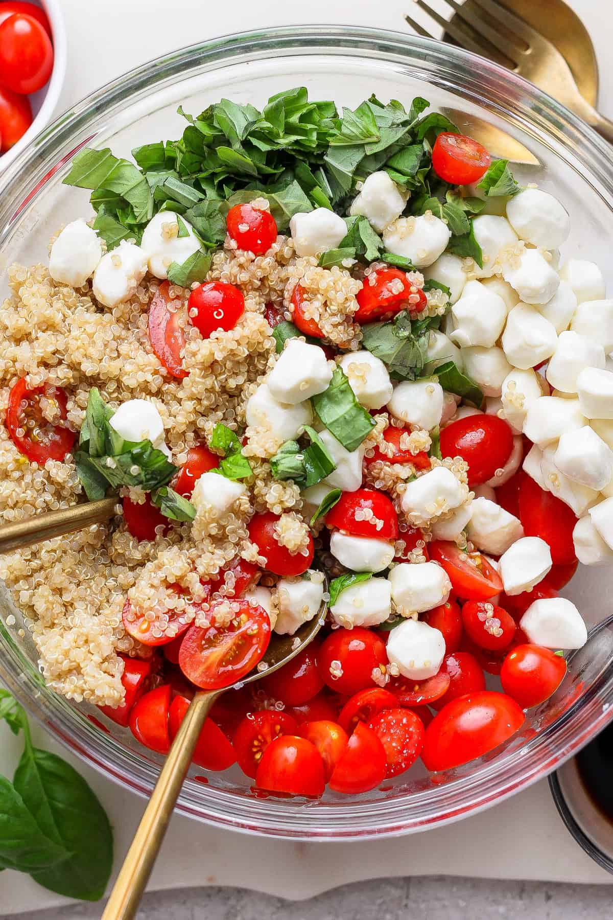 A glass bowl filled with quinoa, halved cherry tomatoes, mozzarella balls, and chopped basil, with two spoons inside.