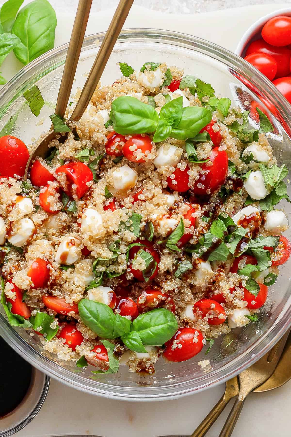 Glass bowl filled with quinoa, cherry tomatoes, mozzarella balls, fresh basil, and balsamic drizzle, with two serving spoons.