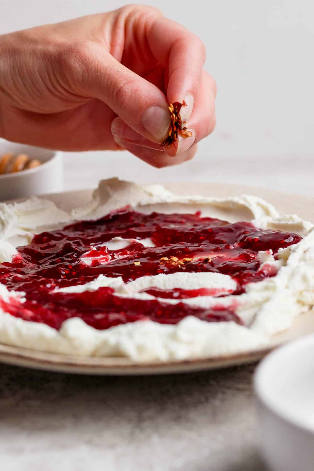 hand sprinkling red pepper flakes over goat cheese dip with raspberry jam