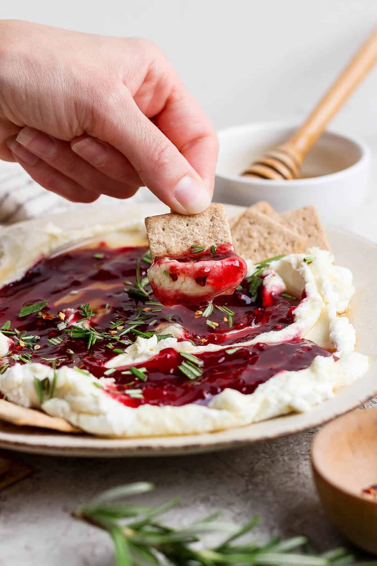 hand lifting a cracker loaded with goat cheese dip and raspberry jam