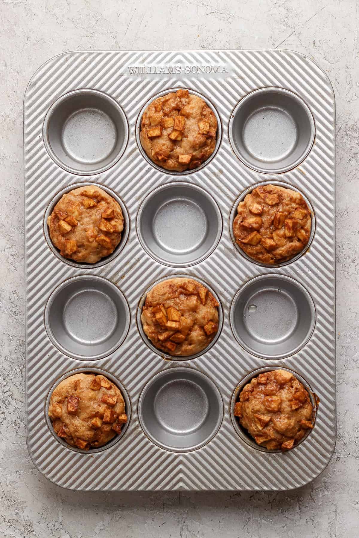 A muffin tin containing six baked muffins with diced apple pieces, with three empty spaces in the tray, on a light textured surface.