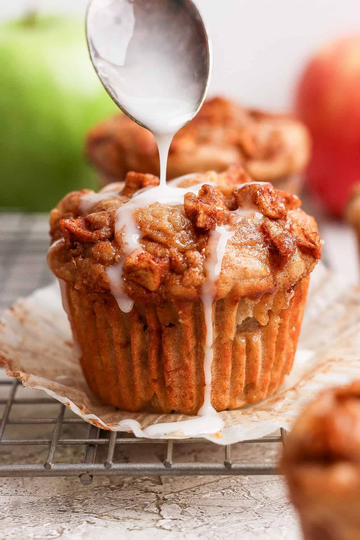 A spoon drizzles white icing over a muffin topped with small pieces of fruit, set on a wire rack with an open paper liner.