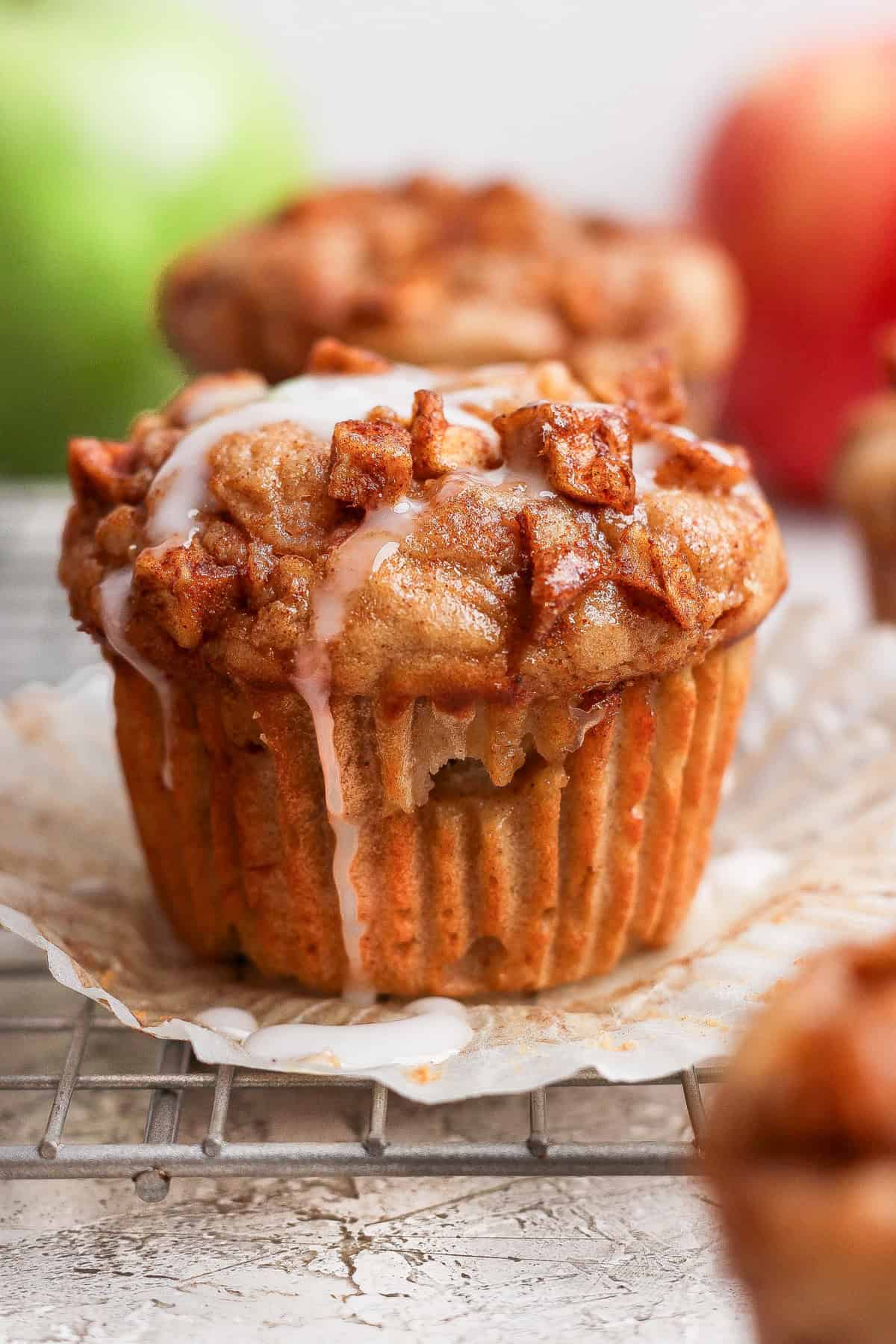 Close-up of an apple cinnamon muffin with icing on top, unwrapped and resting on a cooling rack, with apples blurred in the background.