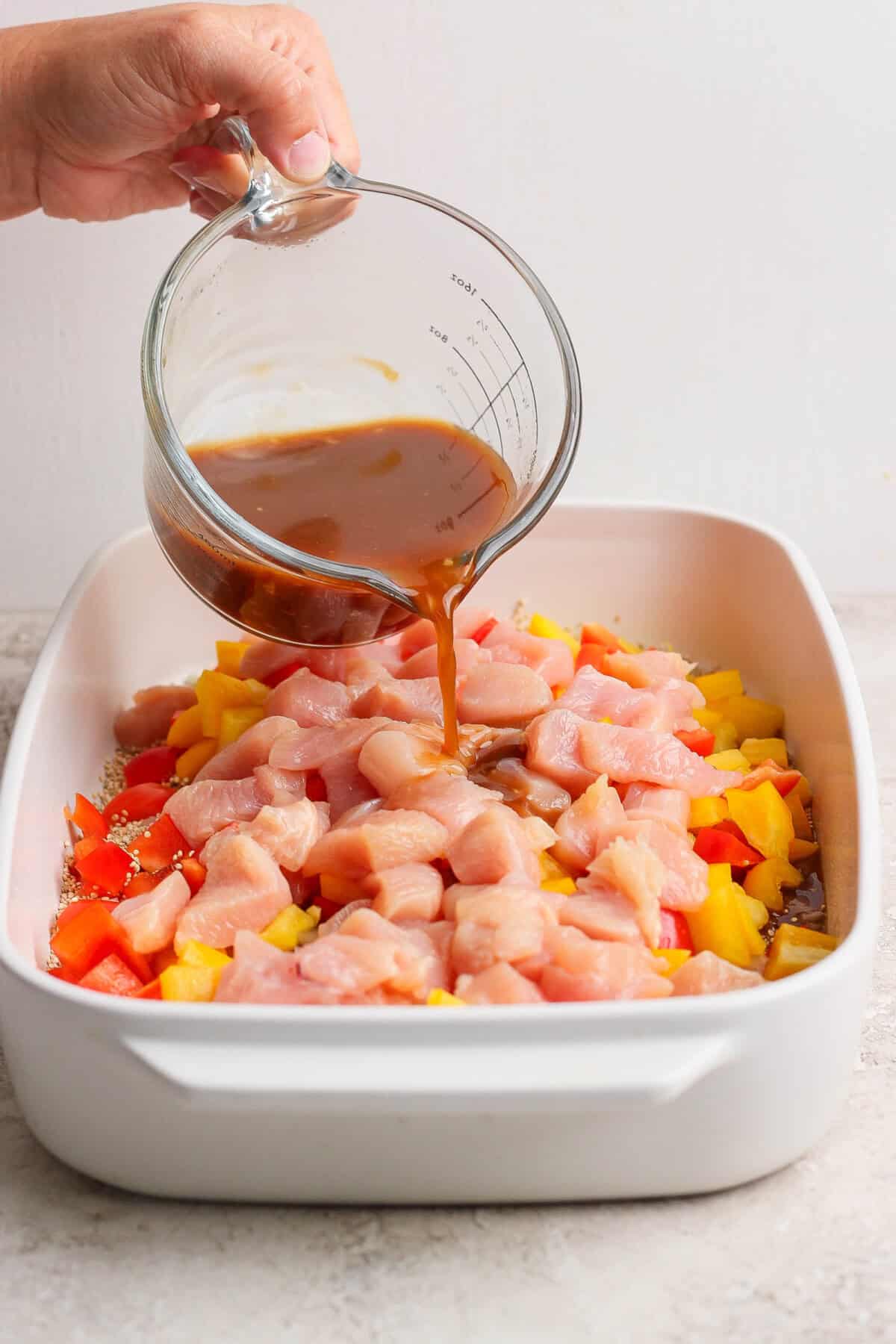 Pouring cashew chicken sauce over raw chicken and bell peppers in a casserole dish.