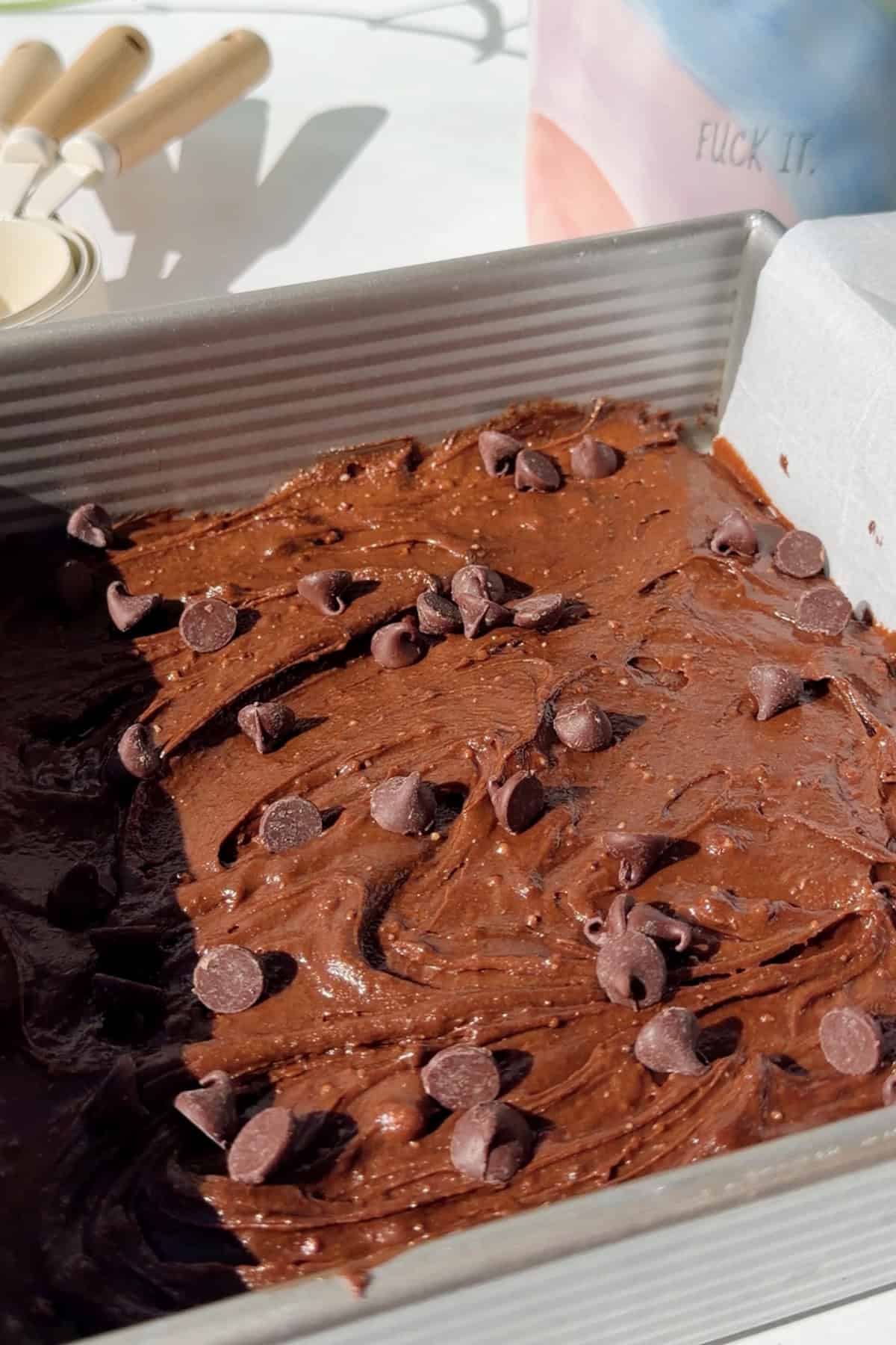 Thick cottage cheese brownie batter spread in a square metal baking pan and topped with chocolate chips before baking.