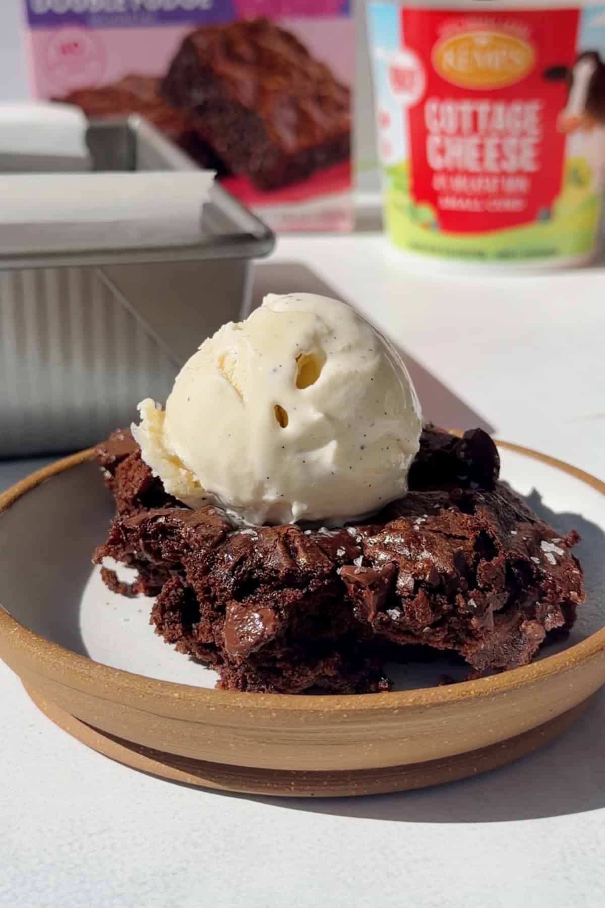 Cottage cheese brownie topped with vanilla ice cream, with the brownie pan and a cottage cheese container in the background.