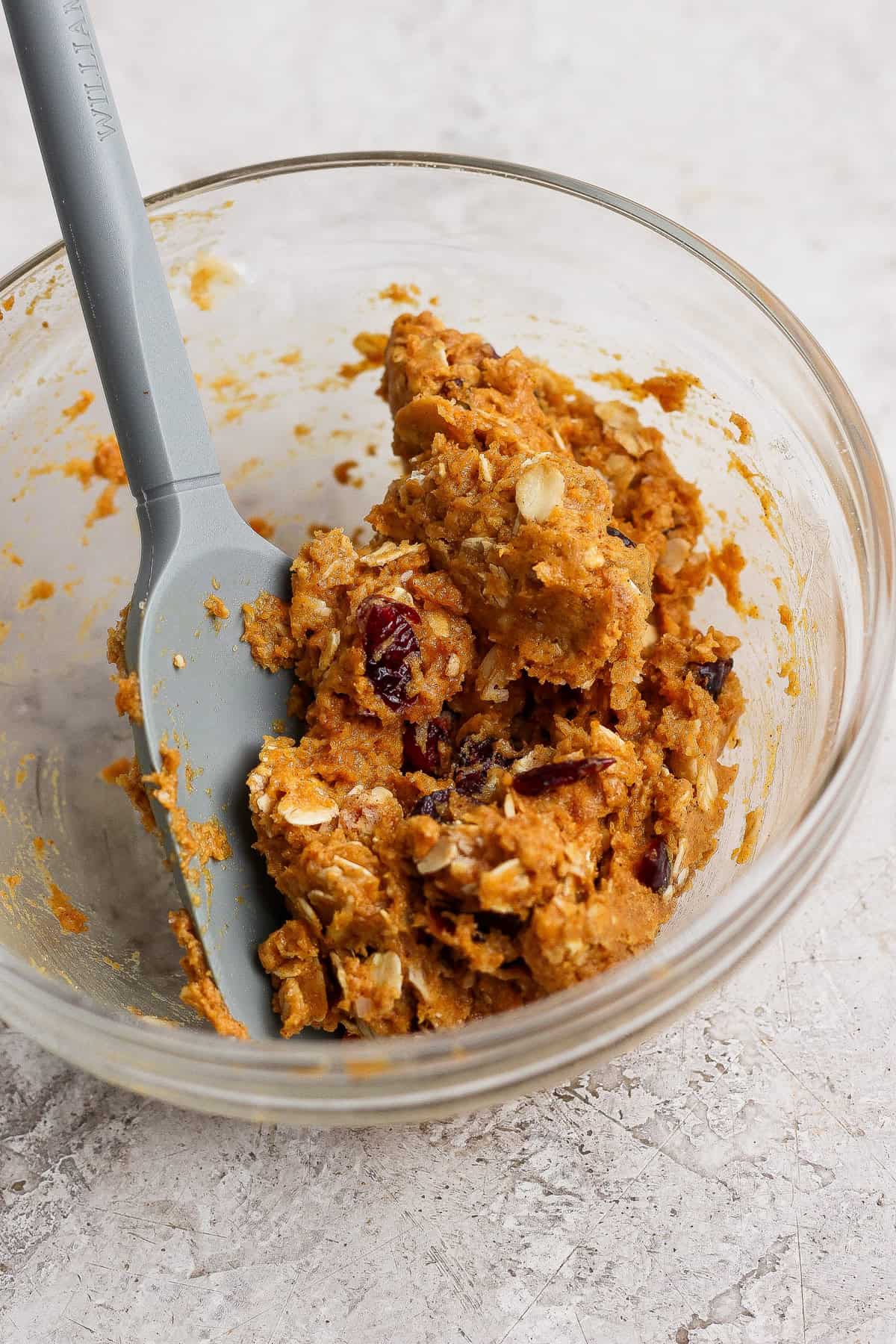 Pumpkin oatmeal cookie dough with dried cranberries in a glass bowl beside a silicone spatula.