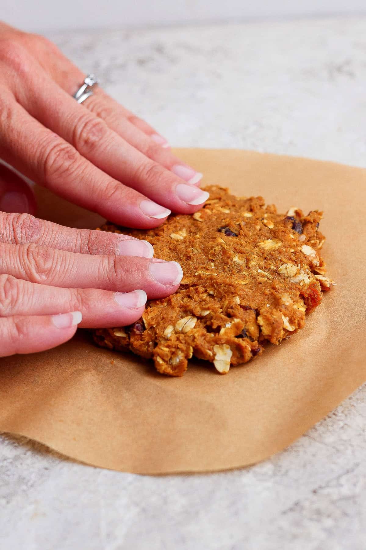 A hand flattens pumpkin oatmeal cookie dough on brown parchment paper.
