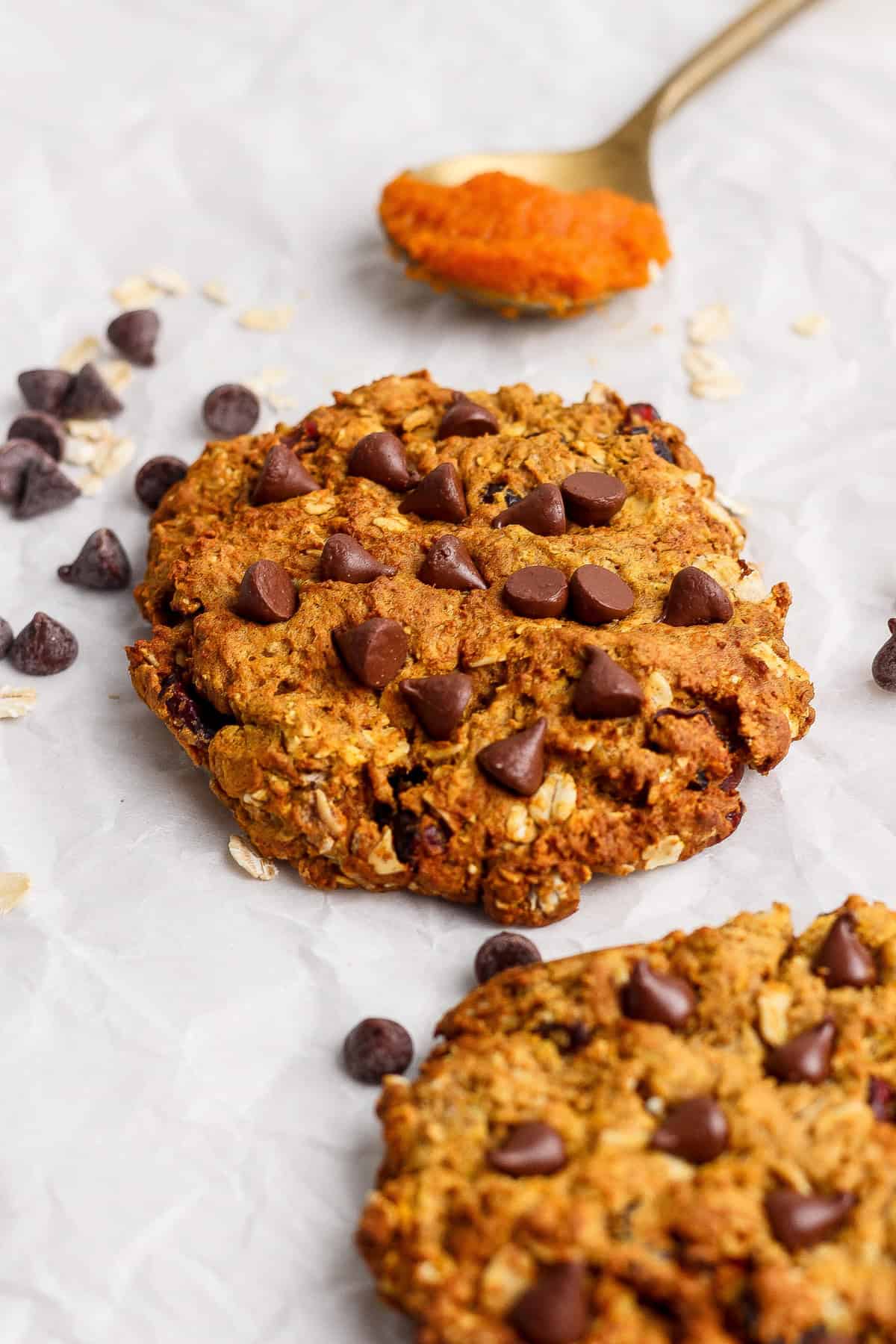 Two pumpkin chocolate chip oatmeal cookies on parchment with scattered oats and chocolate chips.