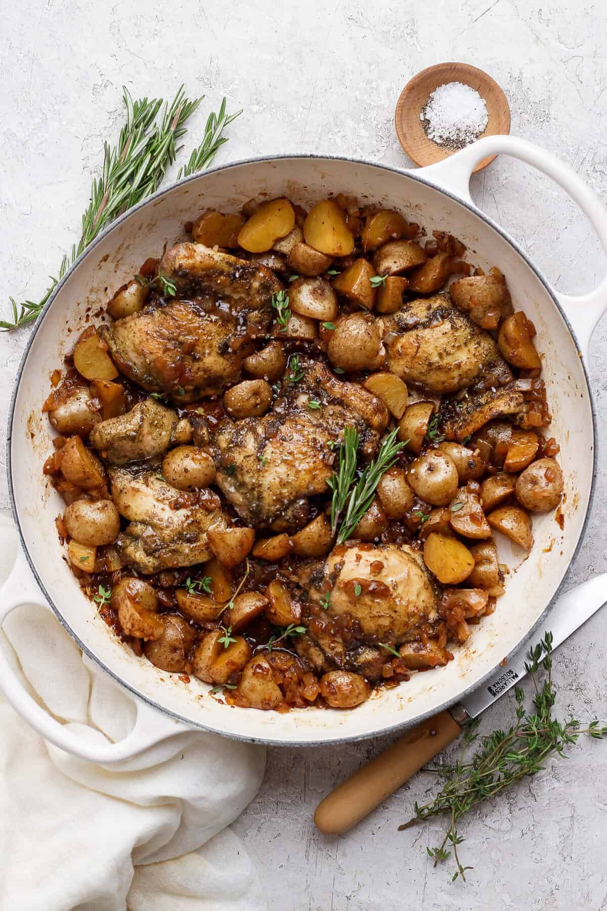 Overhead view of apple cider chicken thighs and baby potatoes in a white skillet with rosemary and thyme.