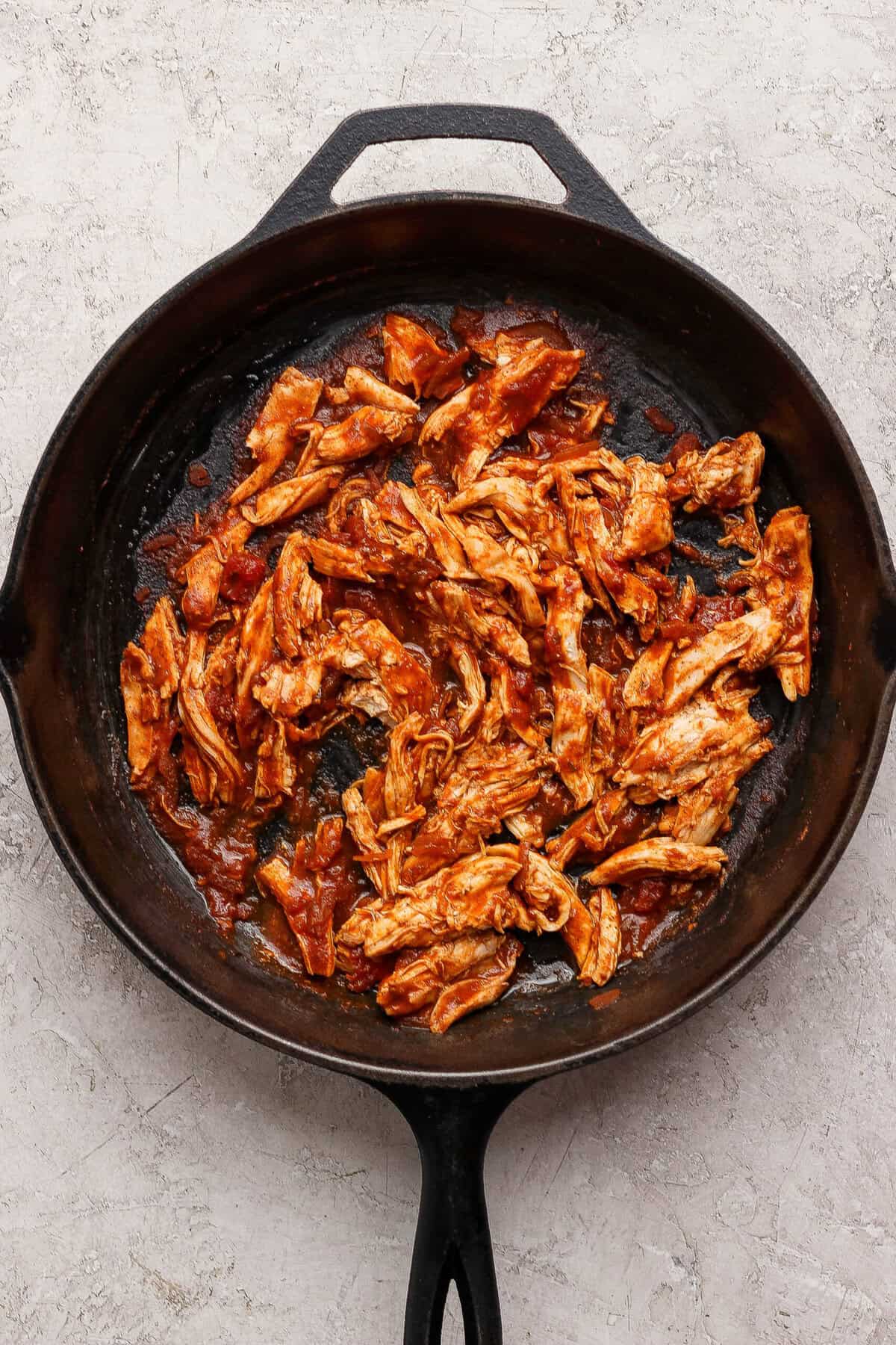 Shredded cooked chicken coated in red taco cooking sauce in a cast-iron skillet.