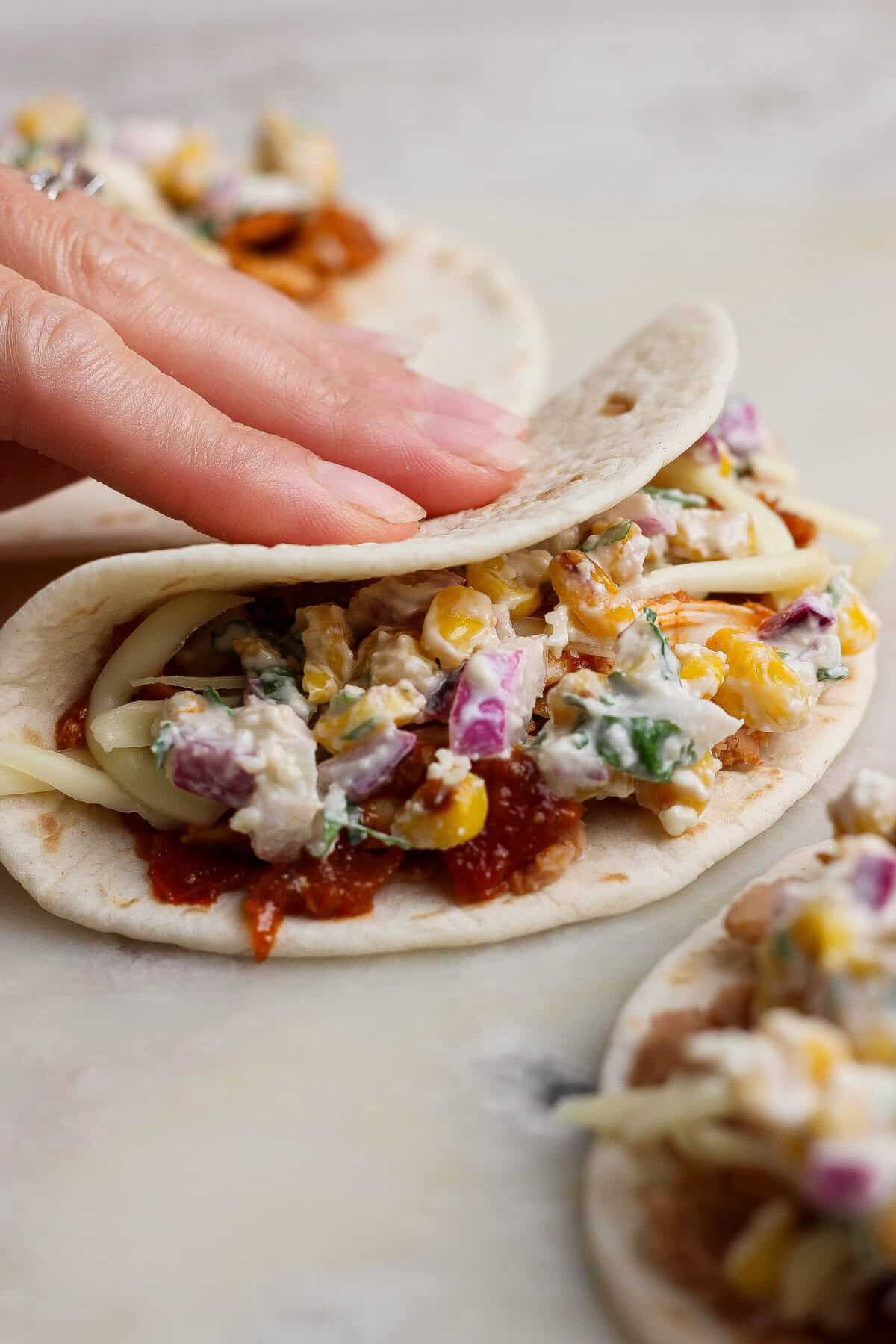 A hand folding a filled flour tortilla over street corn salad, shredded chicken, refried beans, and cheese.