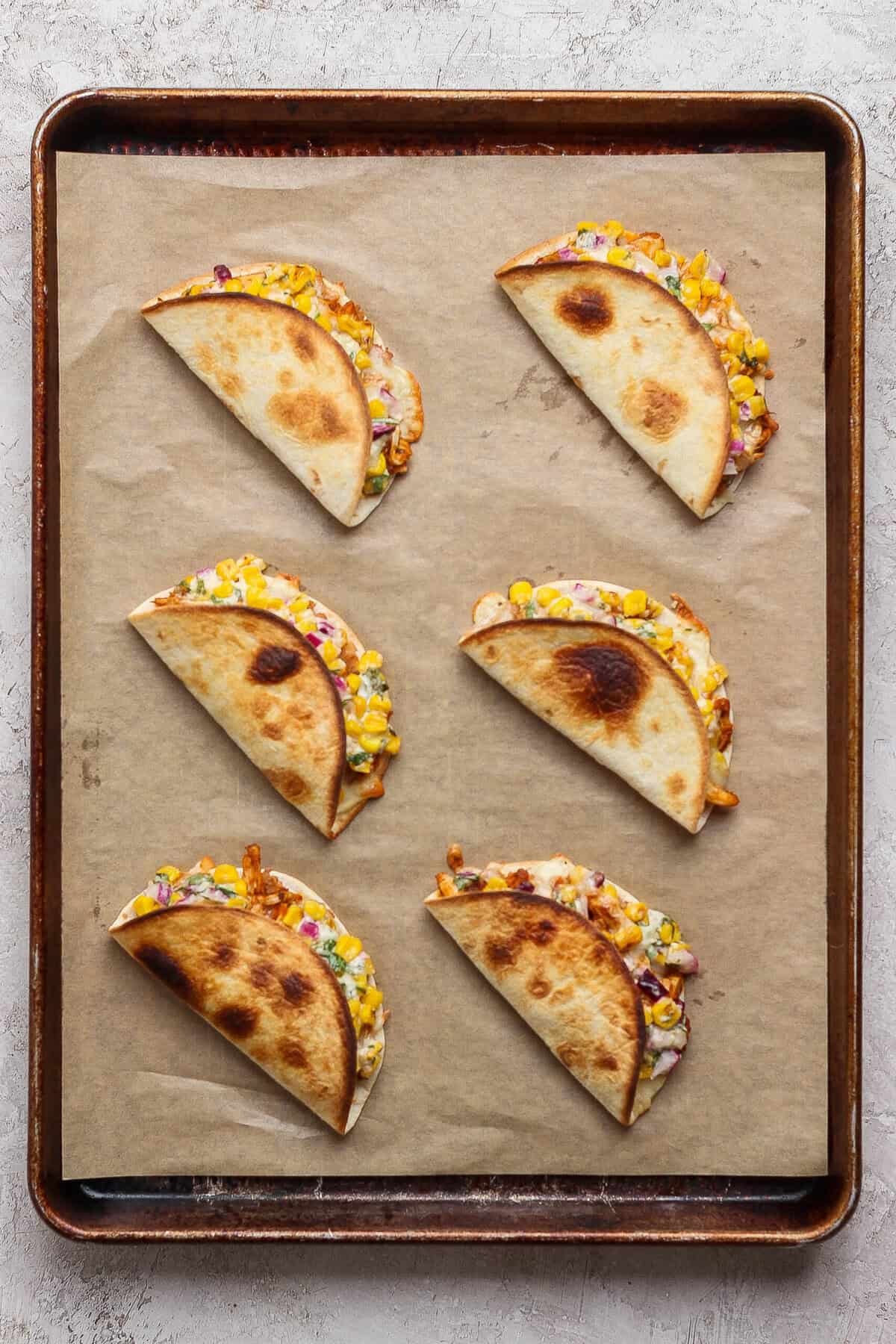 Six folded street corn chicken tacos baked until golden on a parchment-lined metal sheet pan.