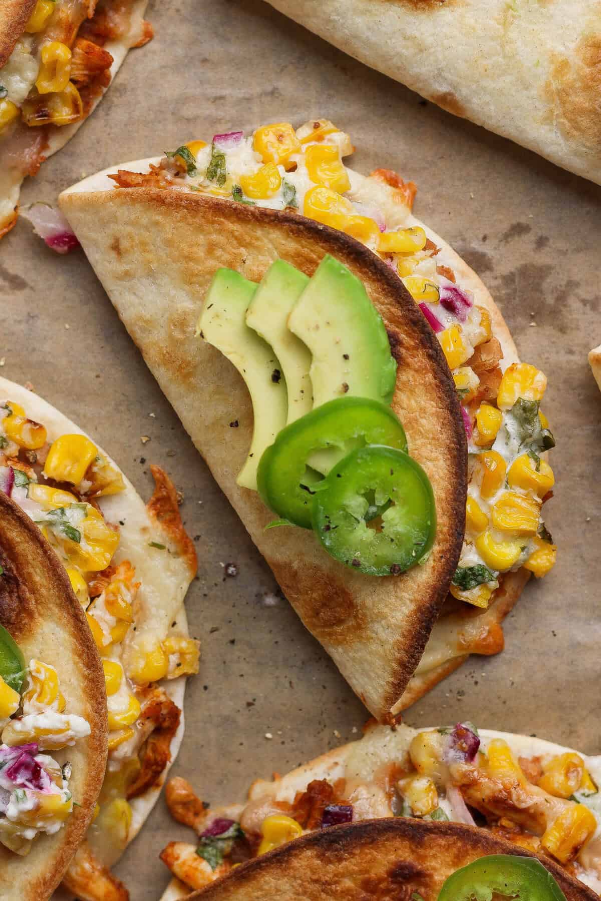 Crispy street corn chicken tacos topped with sliced avocado and jalape&ntilde;o on a parchment-lined baking sheet.